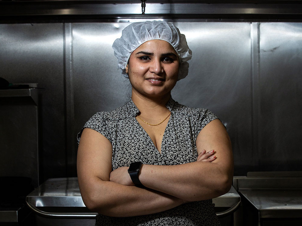 A woman with medium skin tone waring a white hair net stands in a stainless steel kitchen area. She is wearing a short-sleeved patterned blouse, has her arms crossed in front of her and is smiling into the camera.