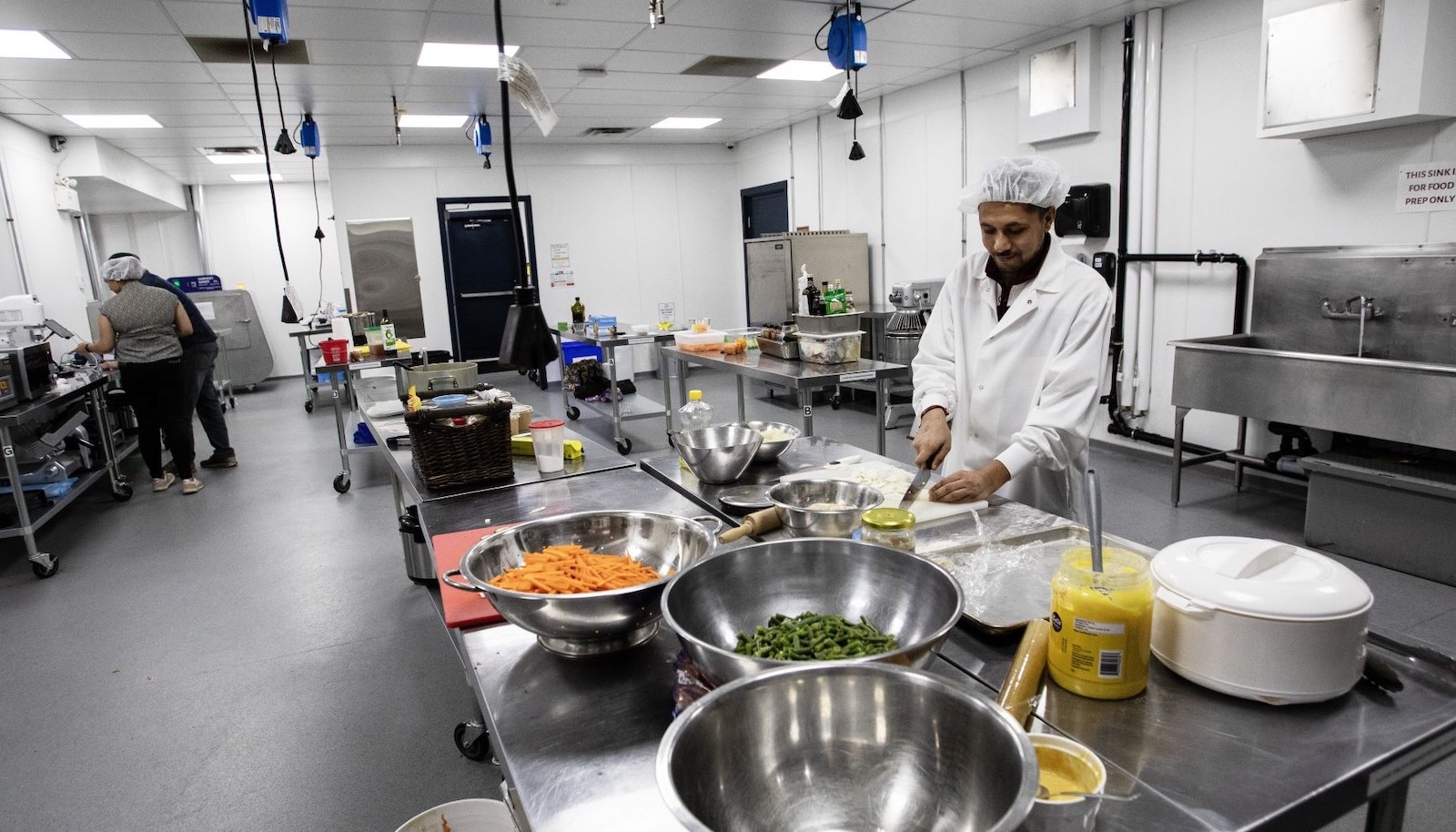 A man with medium skin tone wearing a white hair net and white chef's jacket chops onions on a cutting board surrounded by various prepared vegetables in bowls, in a large kitchen space. At the left of the frame, two people check orders on electronic devices.