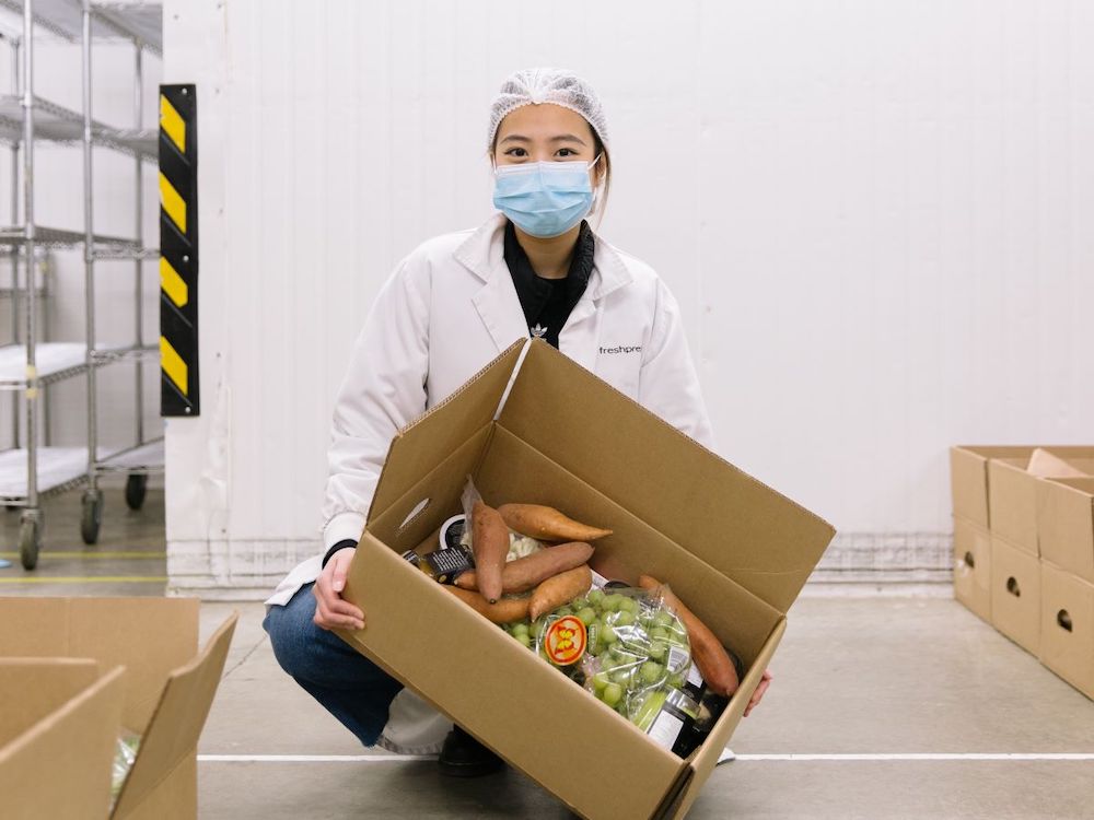 A woman crouches down, wearing a hairnet and mask and holding a cardboard box of produce and other provisions.