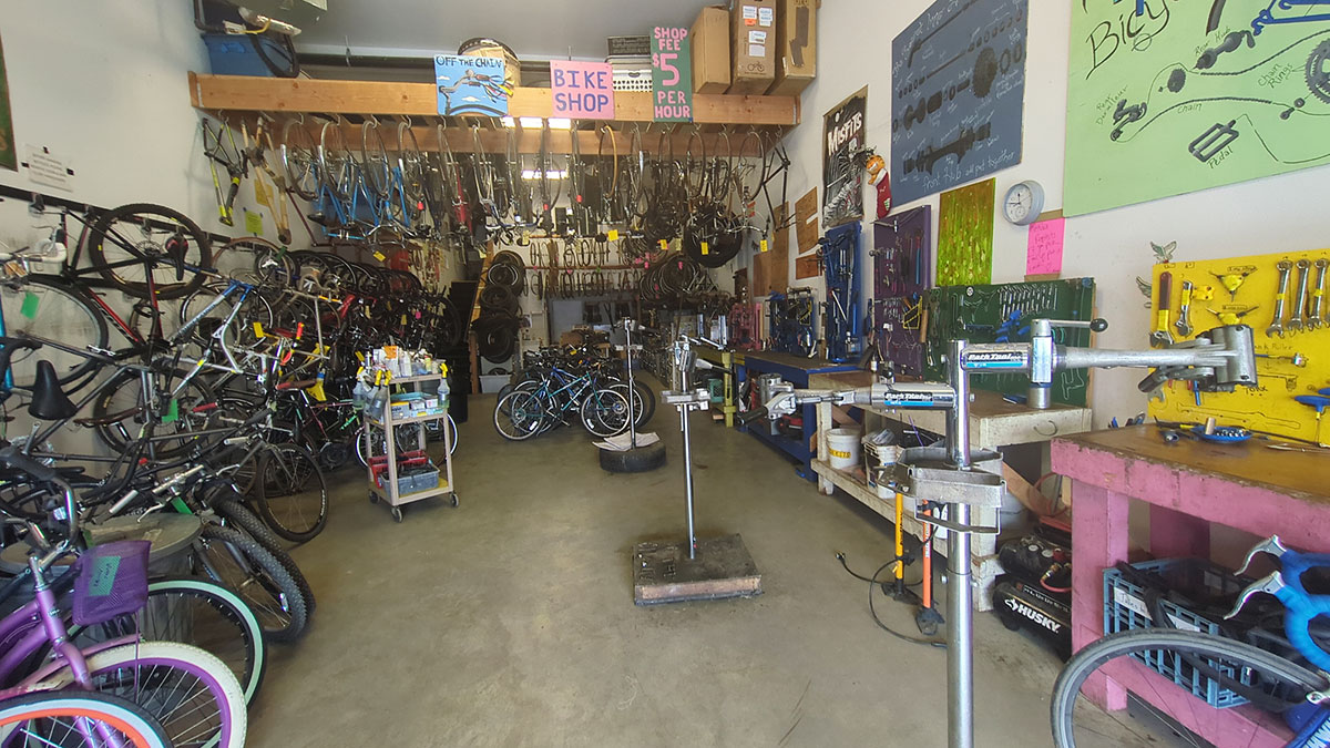A bike shop with a cement floor and white walls has bikes hanging from the ceiling, left wall and on the floor to the left and at the rear of the shop. On the right side are mounted bike tool racks, wooden workbenches and metal bike stands.