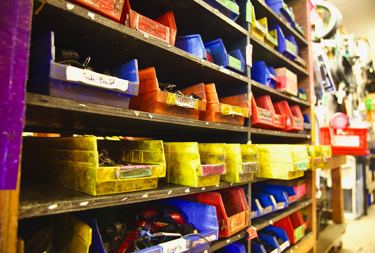 A wooden bookshelf holds rectangular plastic bins in yellow, blue and red. Each bin has a piece of masking tape on the front with the name of the bike part it holds.