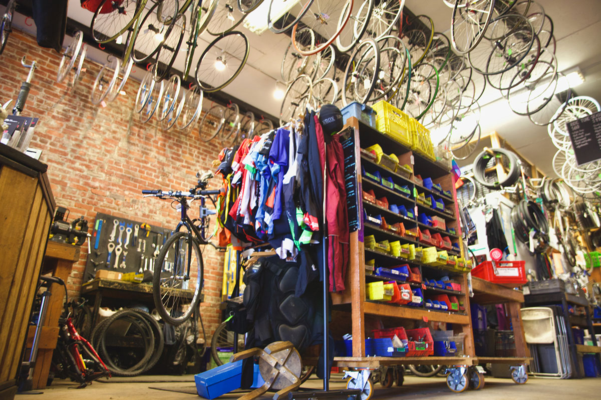 A bike repair shop with wheel frames hanging from the ceiling, and in the middle of the room there are bookshelves on casters holding colourful plastic bins full of sorted used bicycle parts, and a rack of used bicycle clothing.