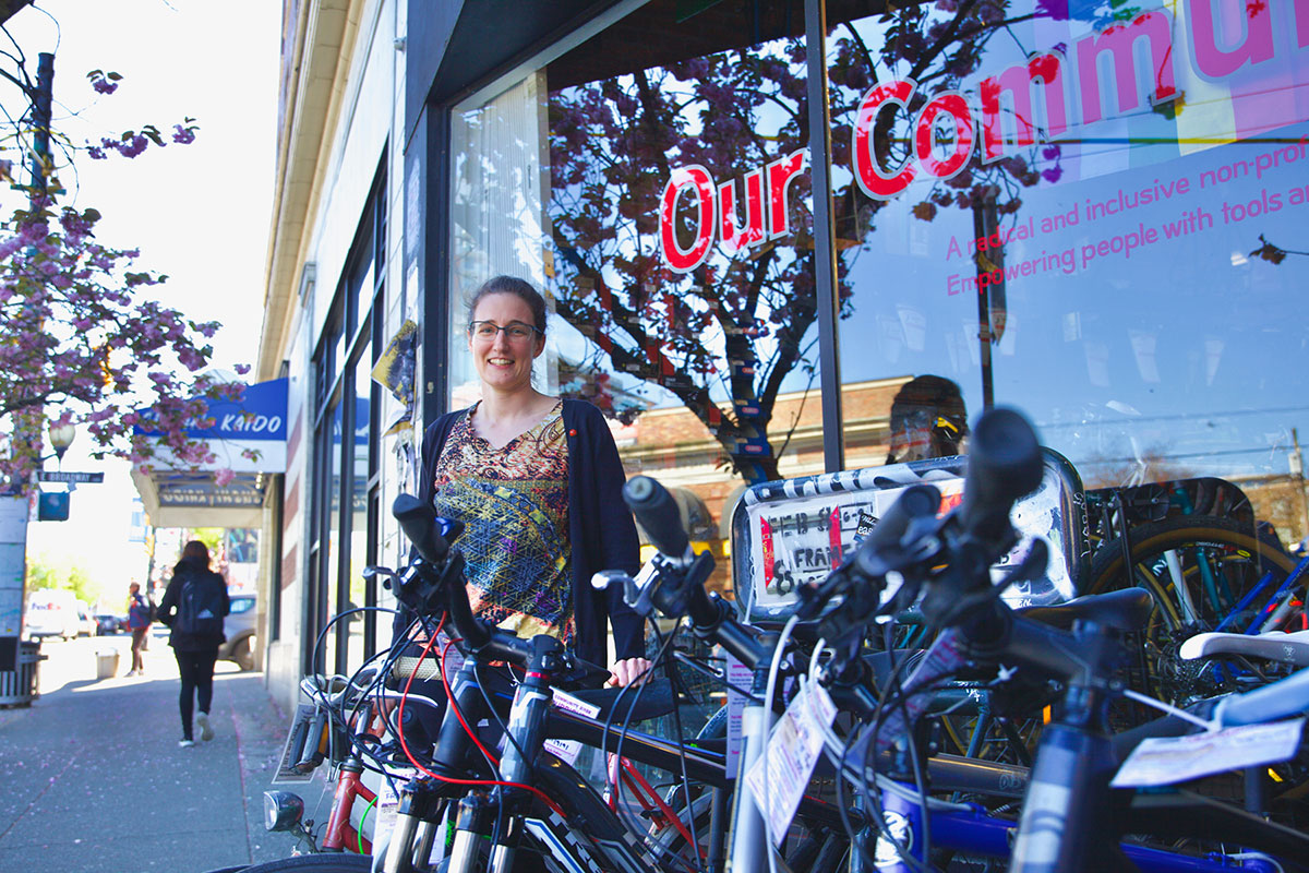 A light-skinned woman stands in front of a storefront window. To the right is a row of bicycles in the foreground, and to the left a sidewalk. The words Our Community Bikes are stencilled in red lettering on the window.