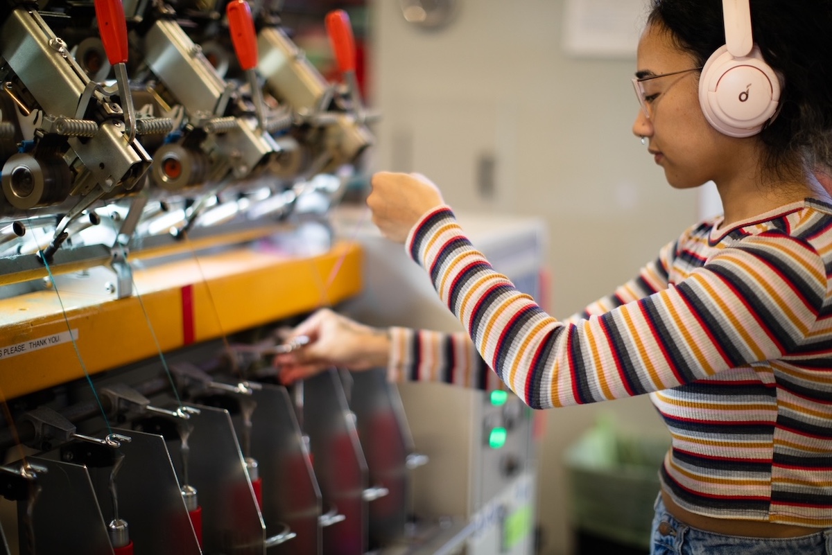 A woman wearing headphones and a striped shirt stretches wool fibre between parts of a machine.