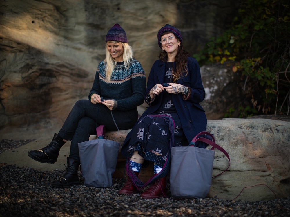 Two women wearing woollen clothing sit outside working on knitting projects. One looks down at what she&rsquo;s knitting. The other smiles at the camera.