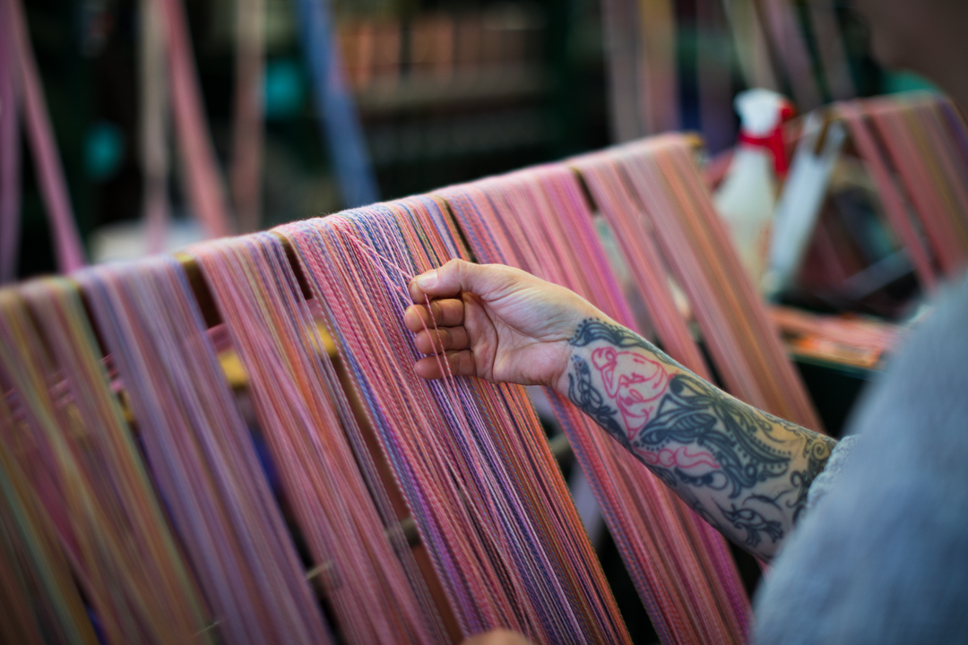 A person with tattooed forearms handles colourful yarn on a machine.