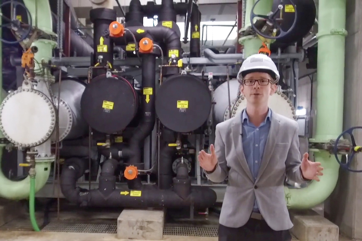 Derek Pope wears a white hard hat, glasses, a light grey visor over a blue button-down shirt and black pants. He has light skin, is gesturing with both hands and is mid-speech. He stands in front of a dense network of thick green, black and white pipes.