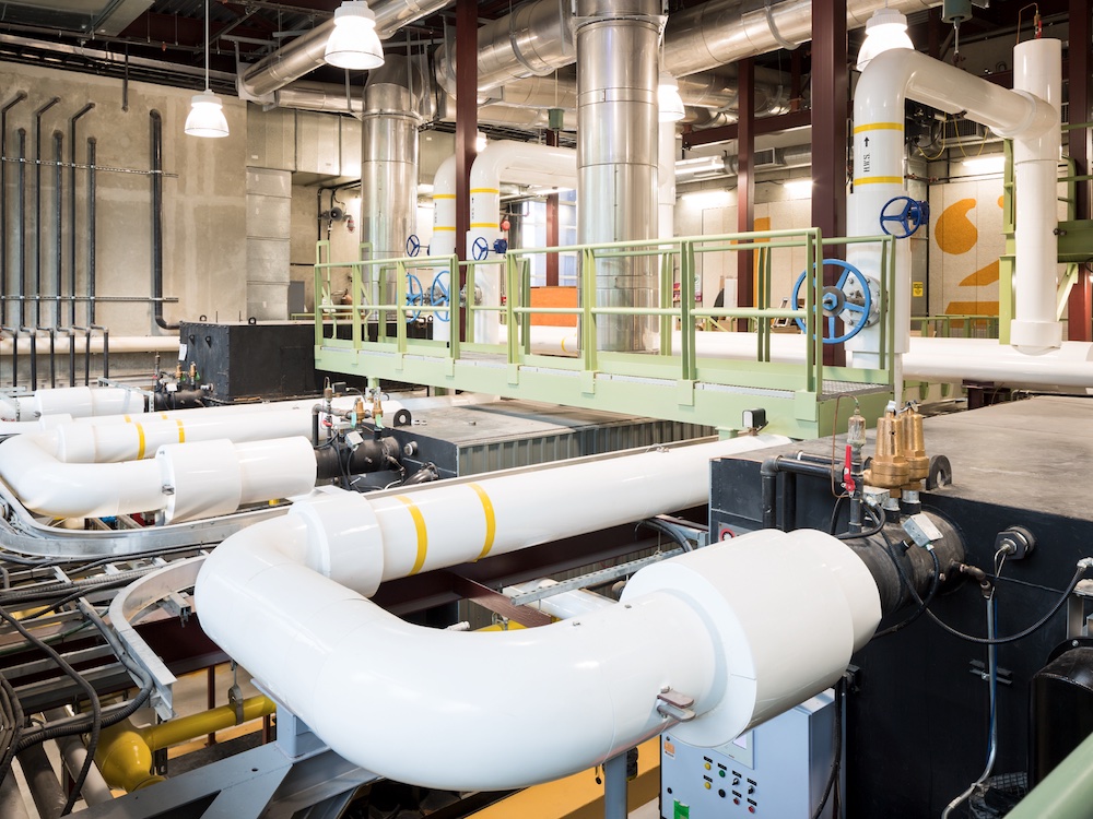 Three sets of thick white pipes with thin yellow stripes are in the foreground of a photograph of the interior of the False Creek Neighbourhood Energy Utility, an industrial facility that recovers heat from raw sewage.