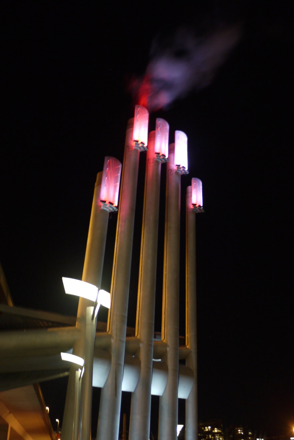 Five tall metal pipes stand outdoors against a dark night sky. The tops of the pipes are lit red, which signifies that more heat is needed for the False Creek Neighbourhood Energy Utility.