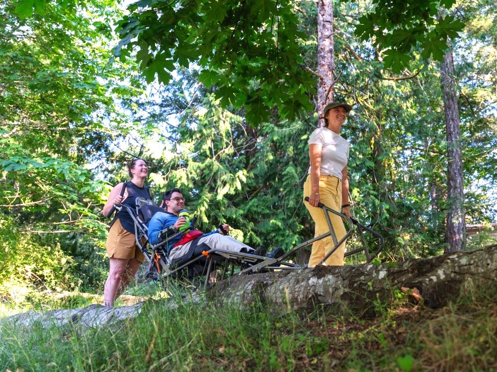 A trio of hikers travel down a leafy path, one riding in a sled-like vehicle designed for outdoor travel by people with disabilities.