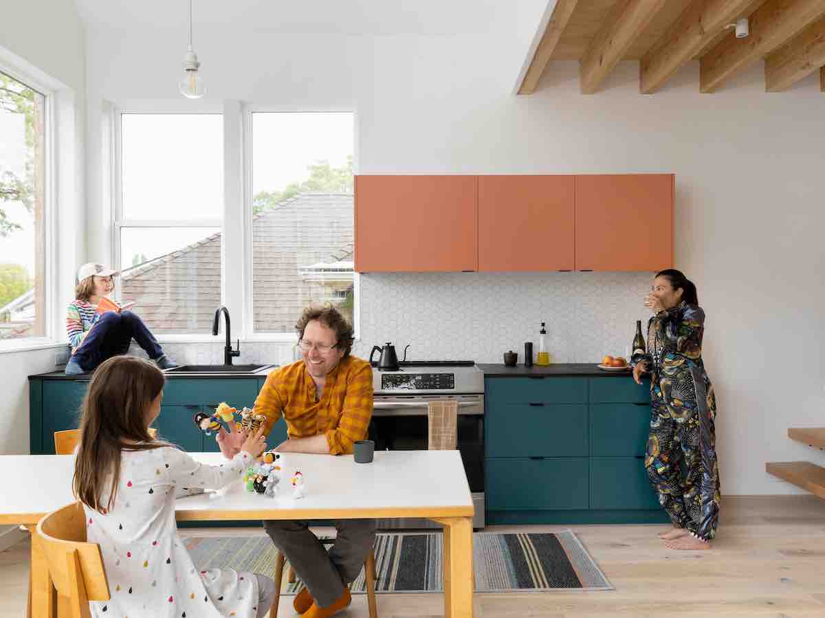A family of four gathers in separate quadrants of a contemporary open-concept kitchen space in a townhome. The cabinets are terra cotta and dark blue, and the walls are white.