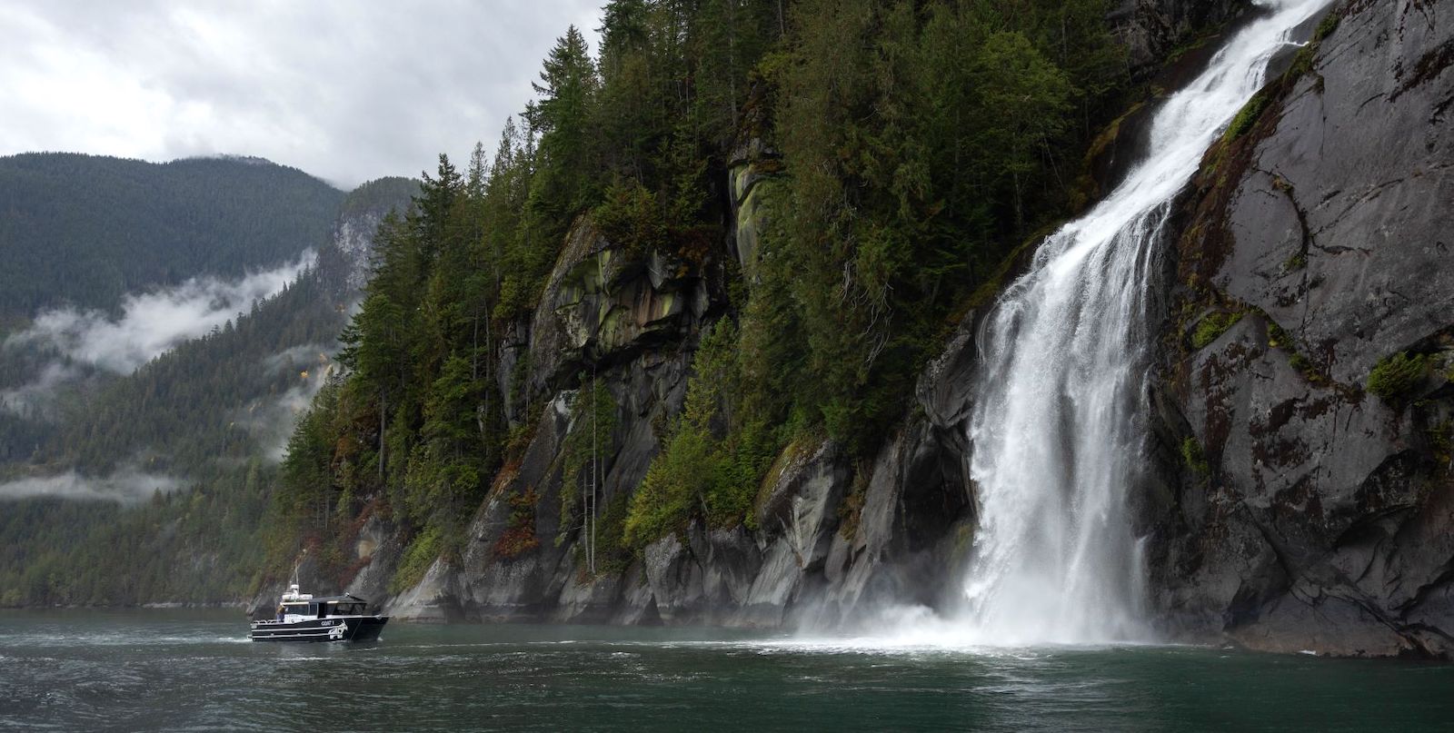 A boat approaches a waterfall cascading into the ocean. In the background are tree-covered mountains dotted with mist.