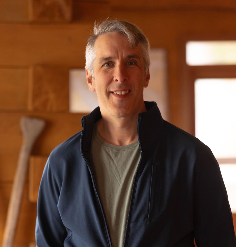 A man with short grey hair and light skin wearing a navy fleece jacket over an olive-green T-shirt smiles at the camera.