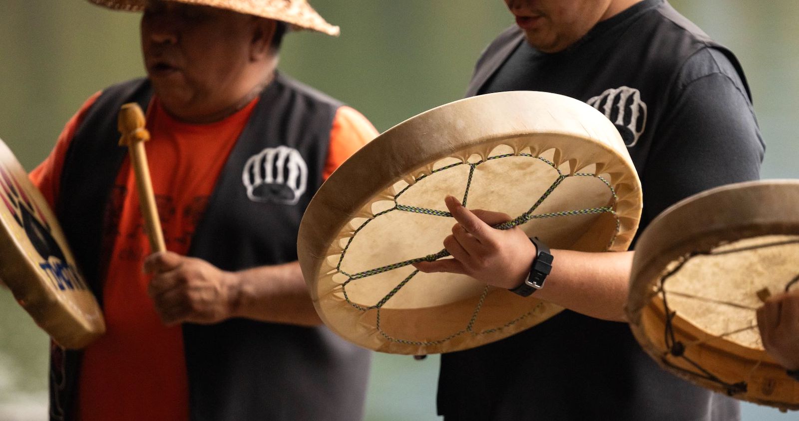 Two people wearing black vests hold Indigenous drums and mallets.