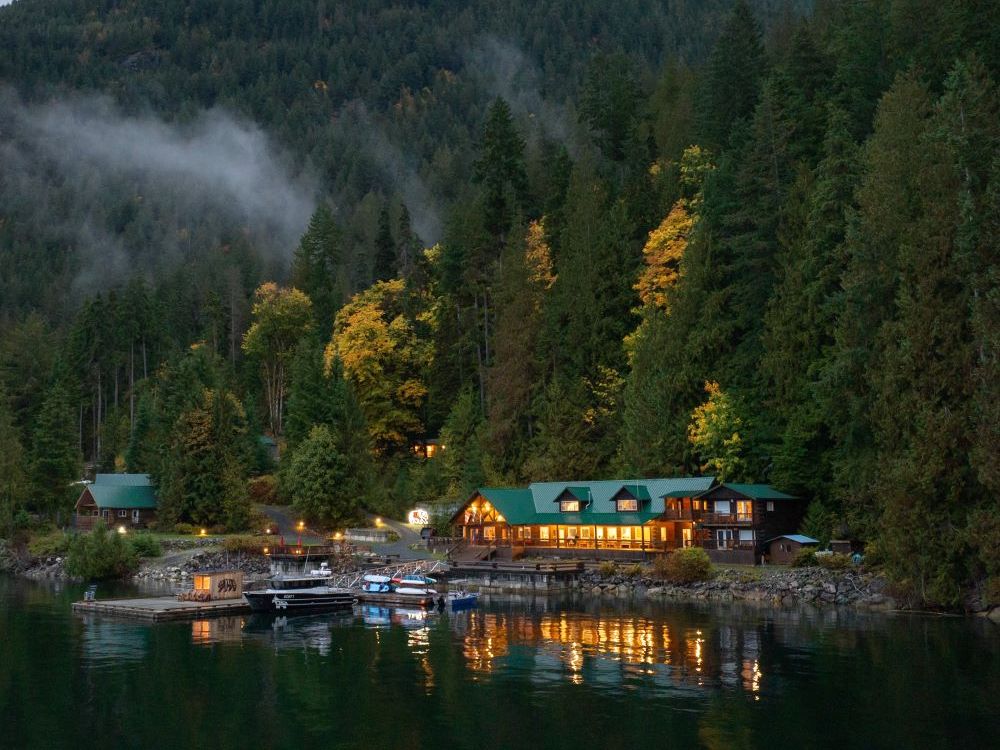 A small waterfront resort, constructed of wood and with a green roof, with windows lit up in the semi-darkness. Immediately behind the building is a hillside covered in dark green conifers interspersed with and a few golden-leaved deciduous trees.