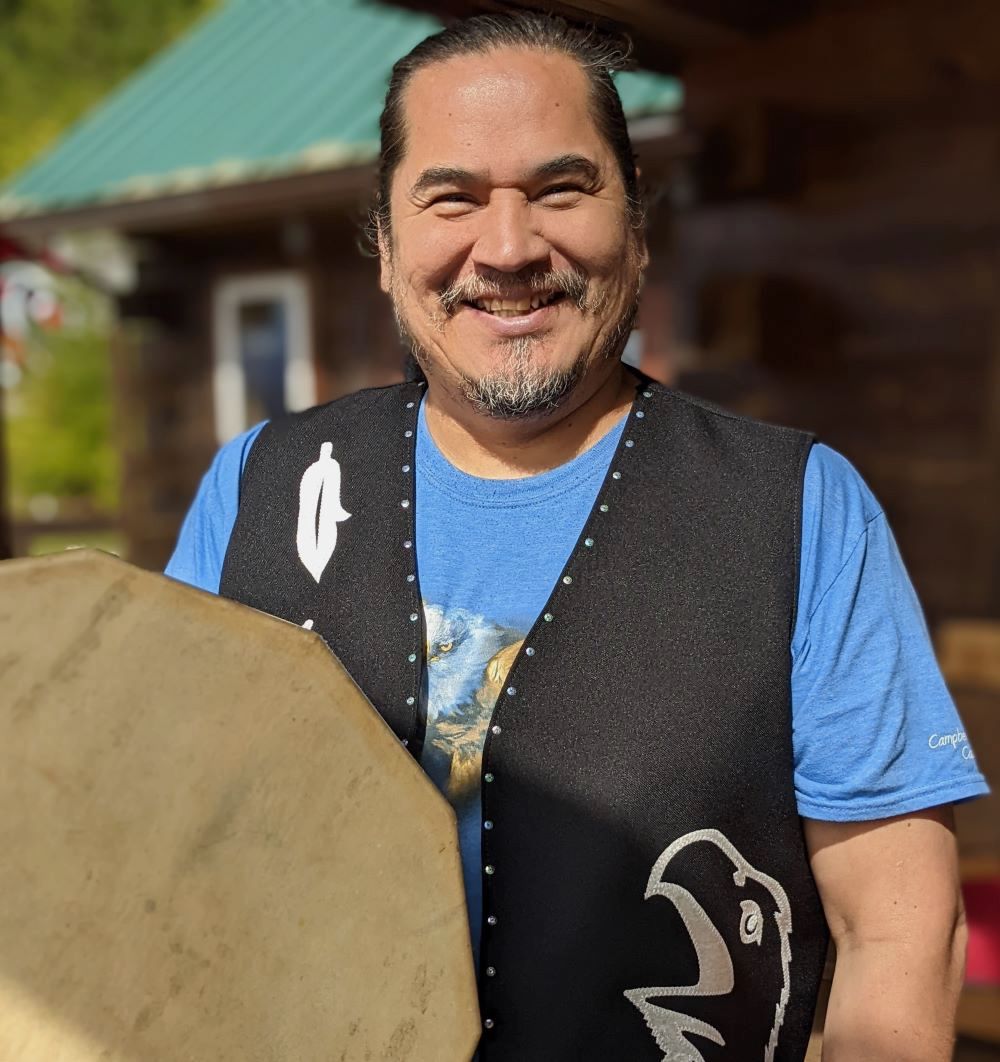 A man with medium-light skin tone and a salt-and-pepper goatee wearing a black vest with white Indigenous designs smiles into the camera.