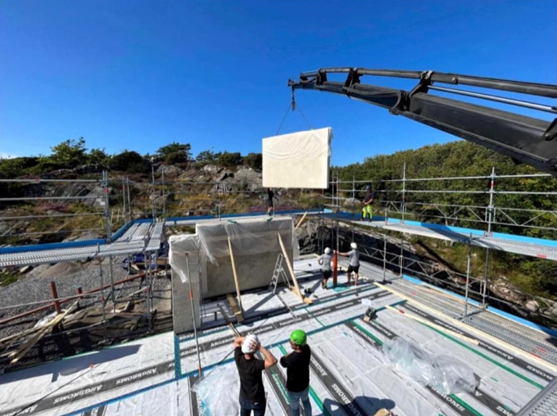 A light beige hempcrete panel is suspended from a crane over a construction site under a bright blue sky.