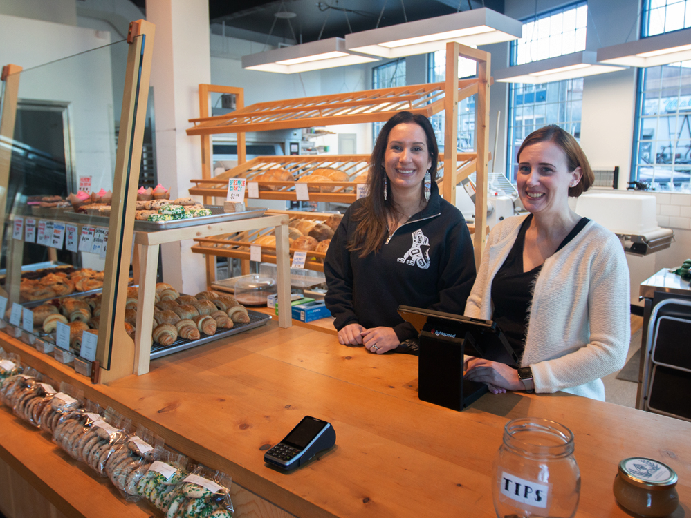 A woman with dark hard wearing a black sweatshirt and long earrings stands next to a woman with fair complexion wearing a white sweater. Behind them next to them is a shelf filled with baked pastries.