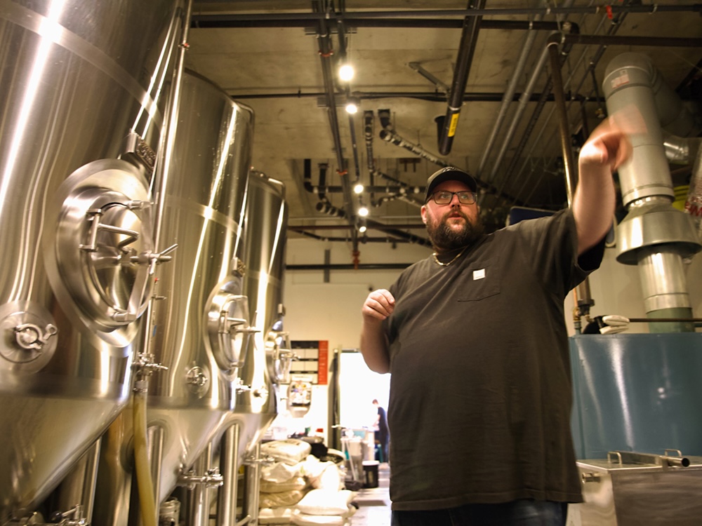 A man with medium-light skin tone wearing a black T-shirt, baseball cap and glasses gestures to something behind the camera. To his left are three large stainless steel vats.