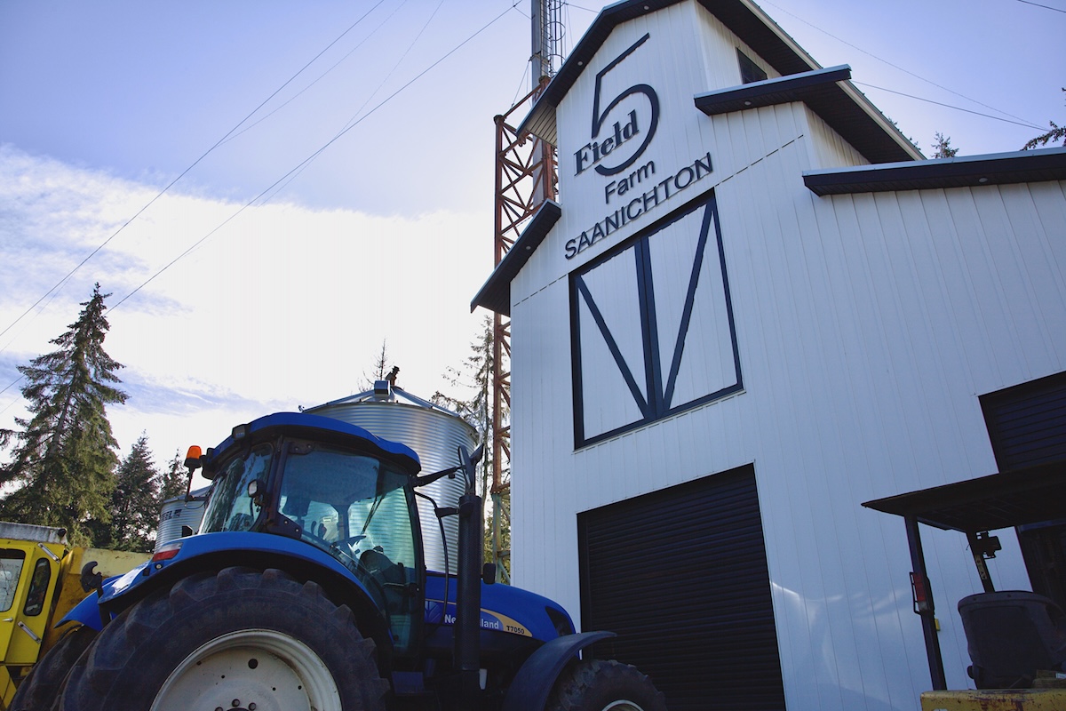 A blue tractor is parked in front of a two-storey white barn with black trim, with the Field 5 Farm logo near the top.