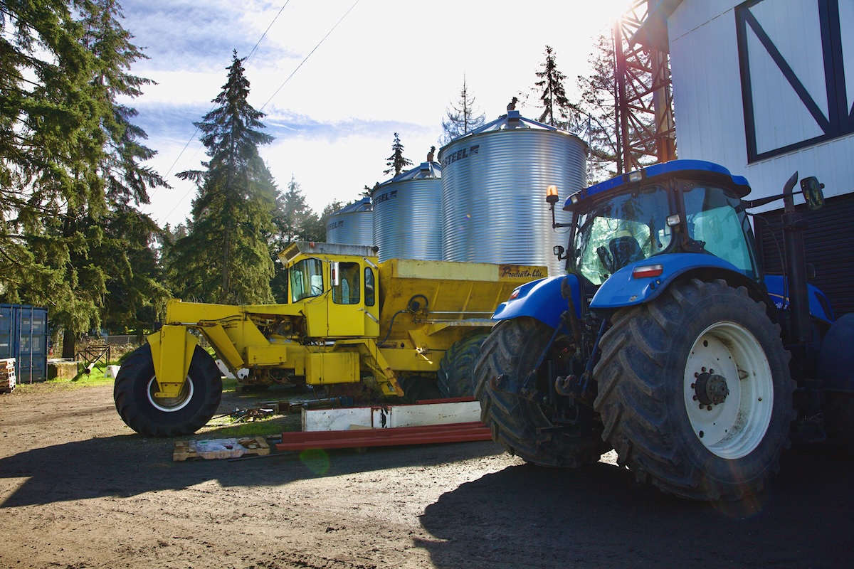 A yellow lime spreader and a blue tractor are parked in front of three silver grain silos.