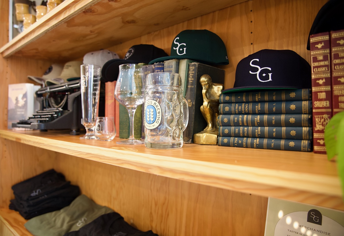 A wooden shelf in a bookcase holds an old typewriter, glassware in front of leather-bound books, and flat-bill baseball hats with an SG logo on the front.