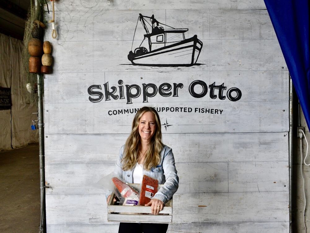A smiling woman with light skin tone and long blond hair holds a wooden crate full of packaged seafood. Behind her is a sign that says &lsquo;Skipper Otto Community-Supported Fishery&rsquo; with an illustration of a small fishing boat.