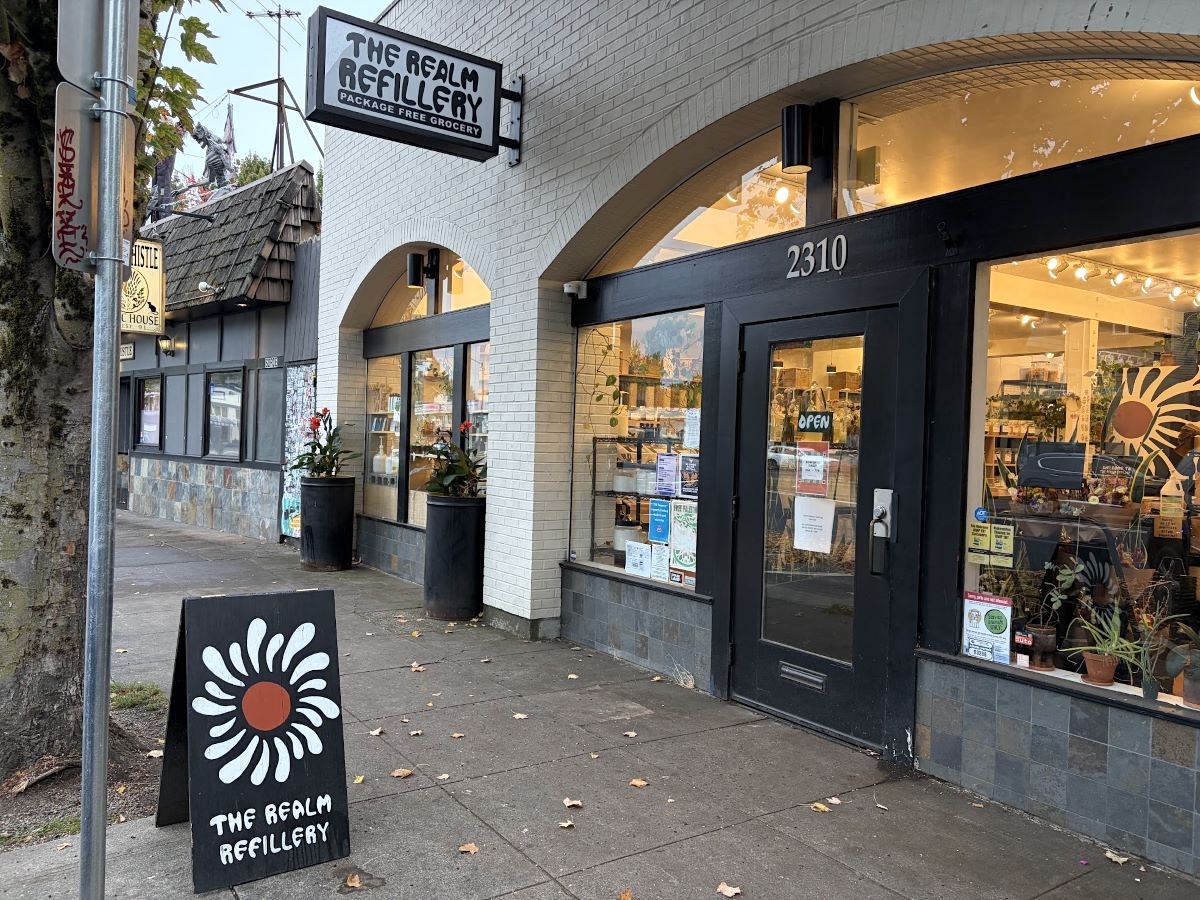 An outside view of a white brick storefront with the words &lsquo;The Realm Refillery&rsquo; on a sign at the top.