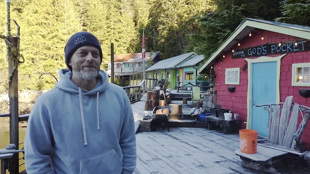 A man in a toque stands on a dock. A building with the words &lsquo;God&rsquo;s Pocket&rsquo; is just over his left shoulder.