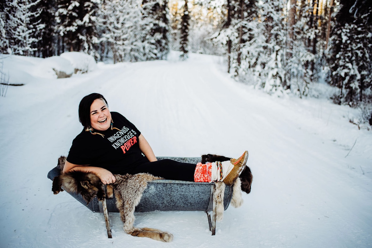A woman sits in the snow, wearing a T-shirt that says, &lsquo;Indigenous Knowledge is Power.&rsquo;