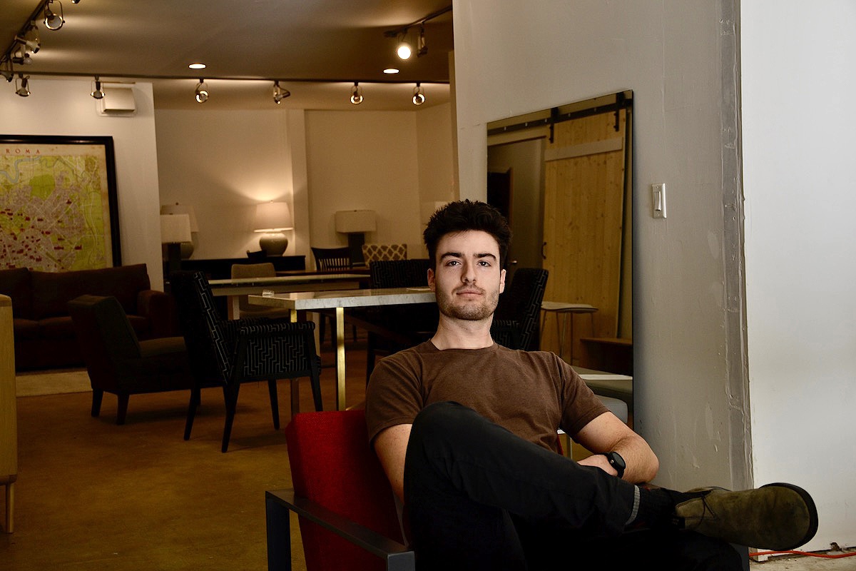 A man in a beige T-shirt sits on a red chair in a warehouse. 