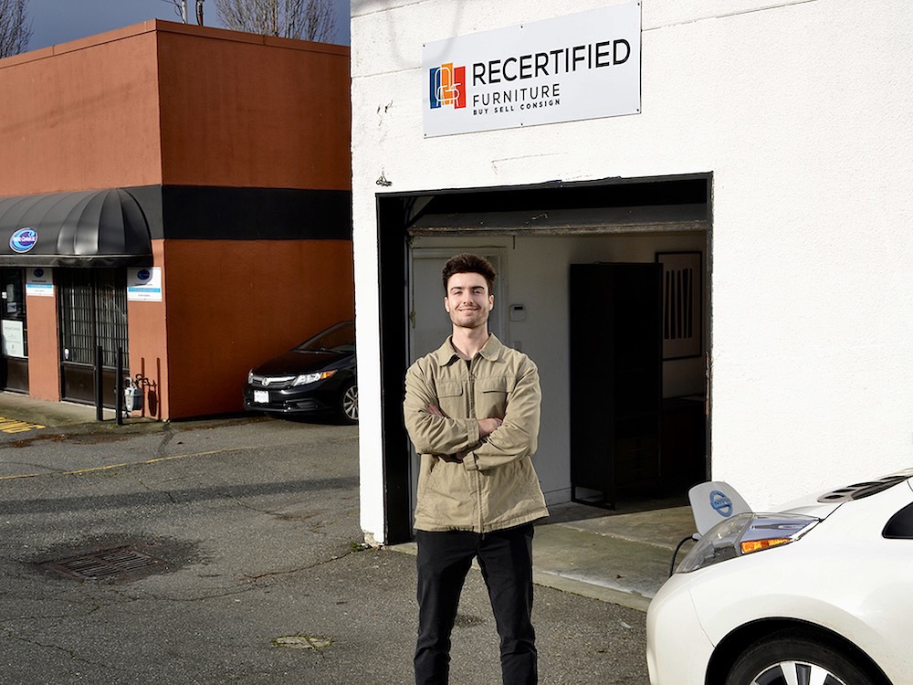 A man in a beige jacket stands with his arms crossed in front of him and smiles for a photo. A sign for Recertified Furniture is posted on a white exterior wall behind him.
