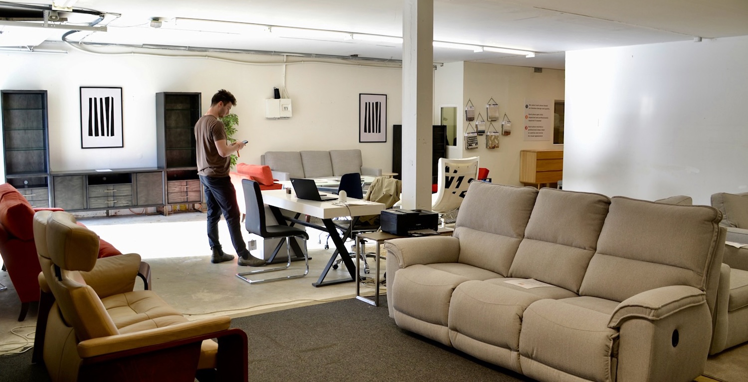 A man on his phone stands in a large warehouse, surrounded by multiple pieces of furniture. 