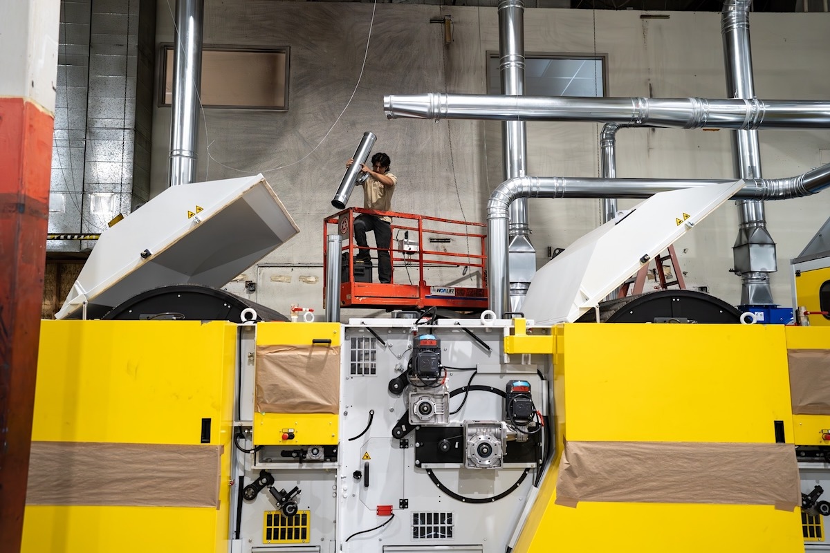 Inside an industrial building, a worker holds a piece of equipment in front of recycling machines.