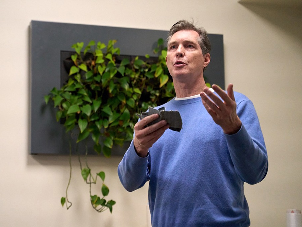 A middle-aged white man in a blue sweater, holding a swatch of fabric, gestures.
