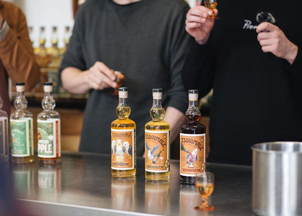 Three multicoloured bottles of Esquimalt Vermouth stand on a table. A pair of bartenders stand behind a table. 