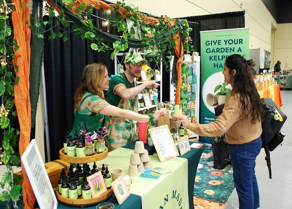 A man and a woman stand in front of a booth at a home and garden show in Seattle. They greet a customer with their hands. Pots, other products and green decorations line their booth. 
