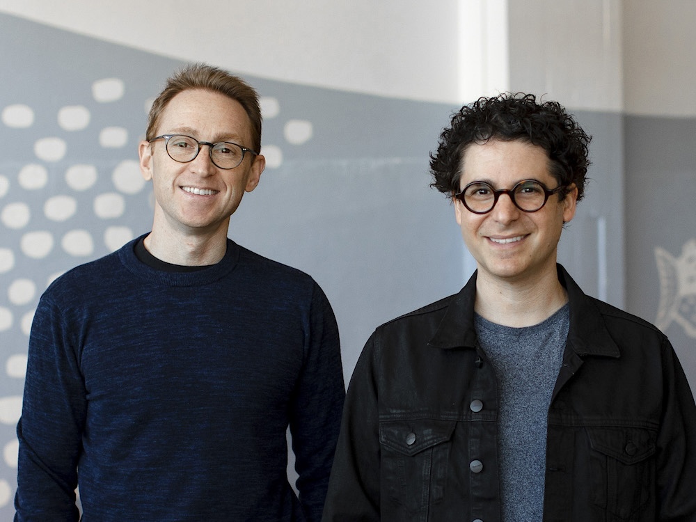 Two men stand side by side in dark shirts. They are both wearing glasses and are standing in front of a blue and white wall.