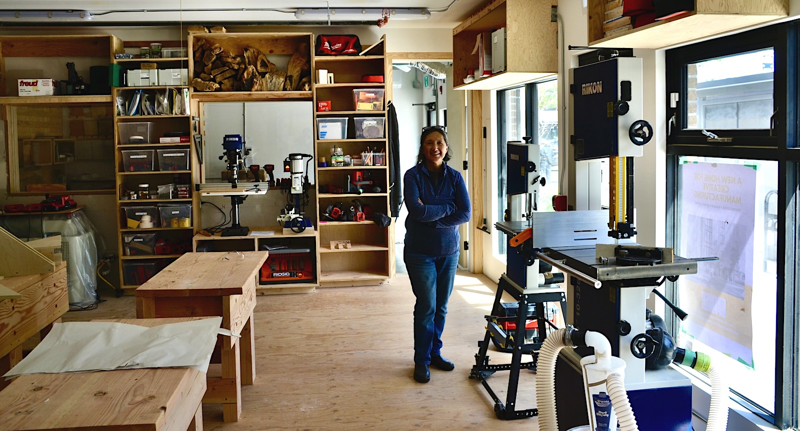 A woman stands in the middle of a wood shop with her arms crossed. There are desks, tools, machines and a variety of equipment in the room. 