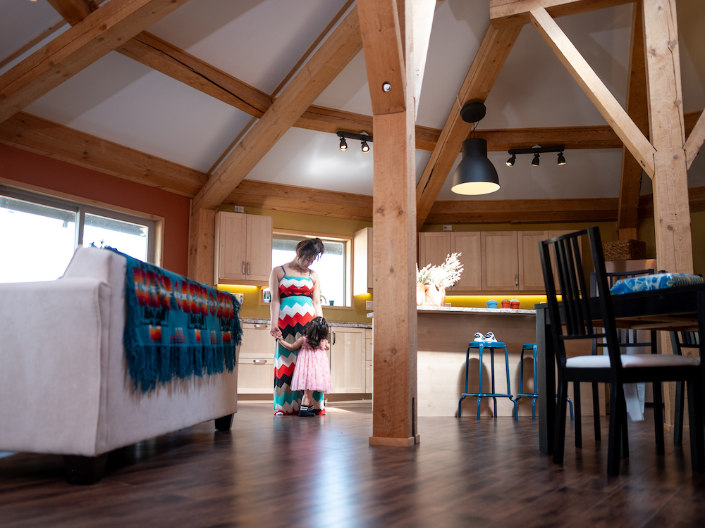 A mother, Lila Rose Zabotel Gott, and daughter, Lyra Averah Rose Zabotel Carlson, embrace in a home built on Skeetchestn territory. The mother is wearing a black, red, white and blue dress while the daughter holds her arms in a pink dress. The kitchen is in the background along with lights and wooden structures. 