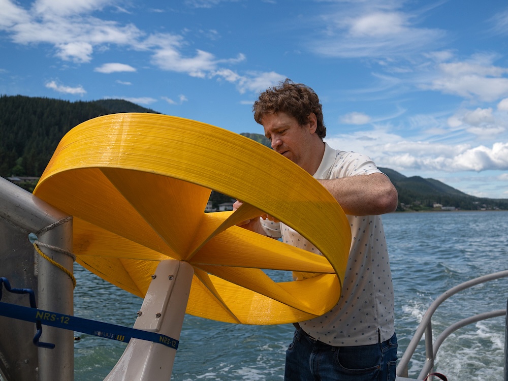 Lance McMullan, dressed in a collared white shirt, works on one of Sitkana&rsquo;s power-generating ocean devices, or PODs, on the back of a boat. The POD is bright yellow. The boat is motoring through an inlet in coastal Alaska.